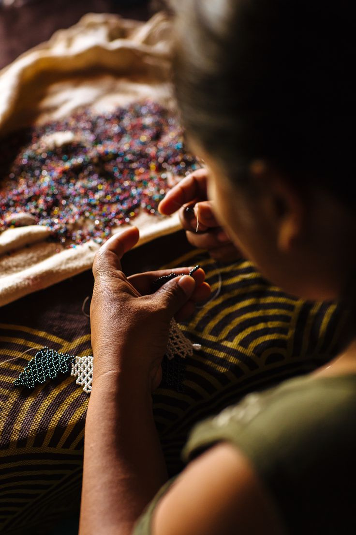 A woman hand-beading by warm light beside a pile of colorful beads — a participant at work.