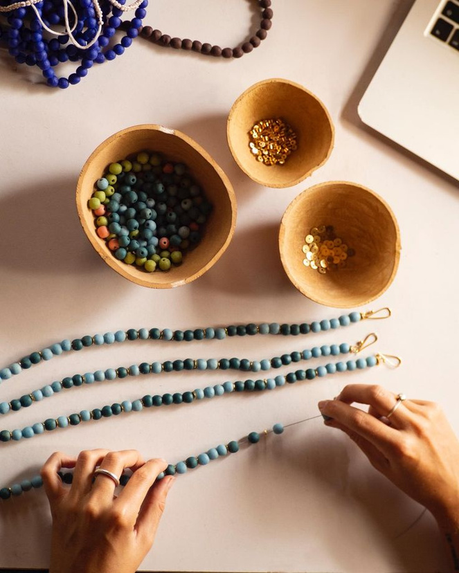 Top-down view of hands stringing turquoise beads beside bowls of beads and sequins — the beading process.
